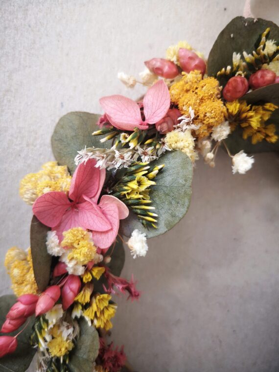 Petite couronne en sarments de vigne ornée de fleurs séchées et d’eucalyptus stabilisé dans des tons printaniers, jaune et rose.