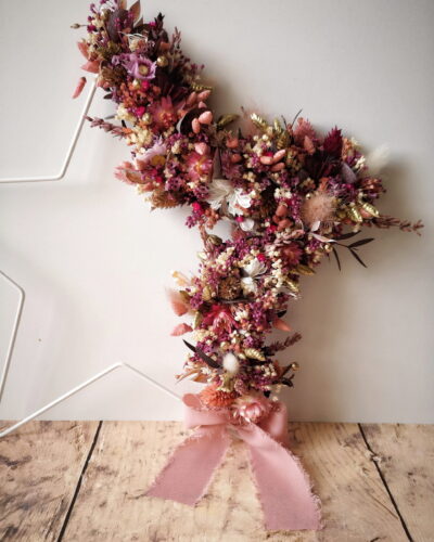 Etoile ornée de fleurs séchées dans les tons rose, doré et violet. Cette couronne en forme d’étoile pourra être suspendue ou posée.