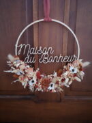 Couronne en bois ornée de fleurs séchées dans les tons terracotta, beige et blanc. Cette couronne en bois fleurie comporte les mots 