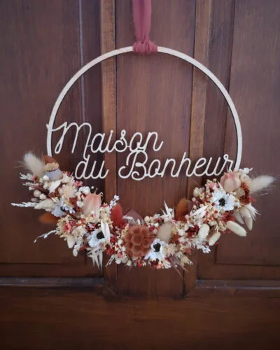 Couronne en bois ornée de fleurs séchées dans les tons terracotta, beige et blanc. Cette couronne en bois fleurie comporte les mots "Maisons du Bonheur".