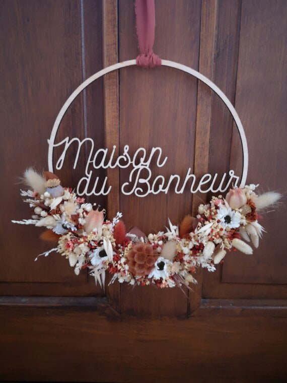 Couronne en bois ornée de fleurs séchées dans les tons terracotta, beige et blanc. Cette couronne en bois fleurie comporte les mots "Maisons du Bonheur".