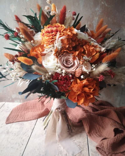 Bouquet de mariage dans les tons terracotta, beige avec du feuillage. Articulé autour d'un dhalia stabilisé, de grosses roses nude et crème et des fleurs dans les teintes rouille, orange et pêche.