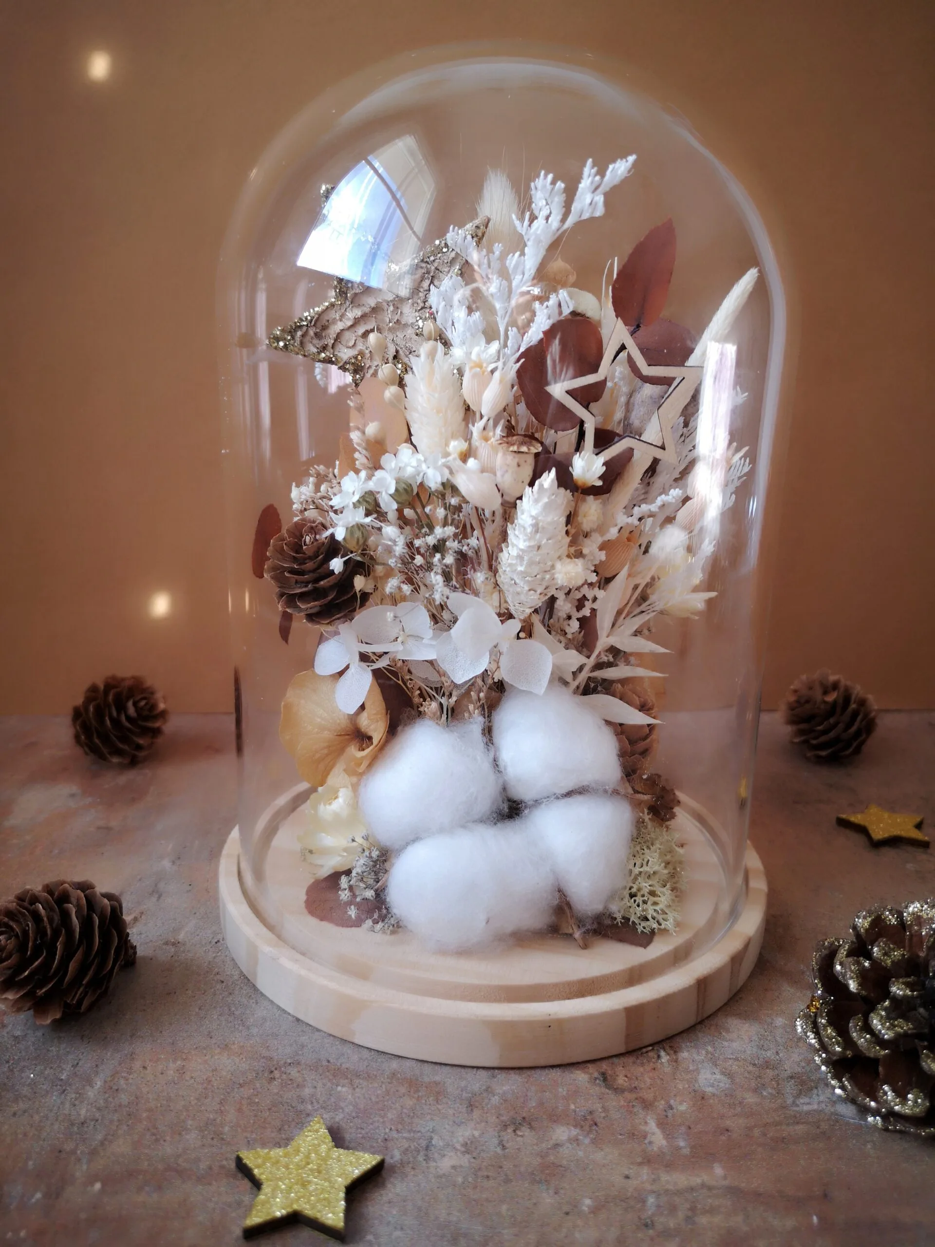 Cloche de fleurs séchées dans des tons naturels (blanc, beige, brun) avec pommes de pin, fleur de coton et étoiles